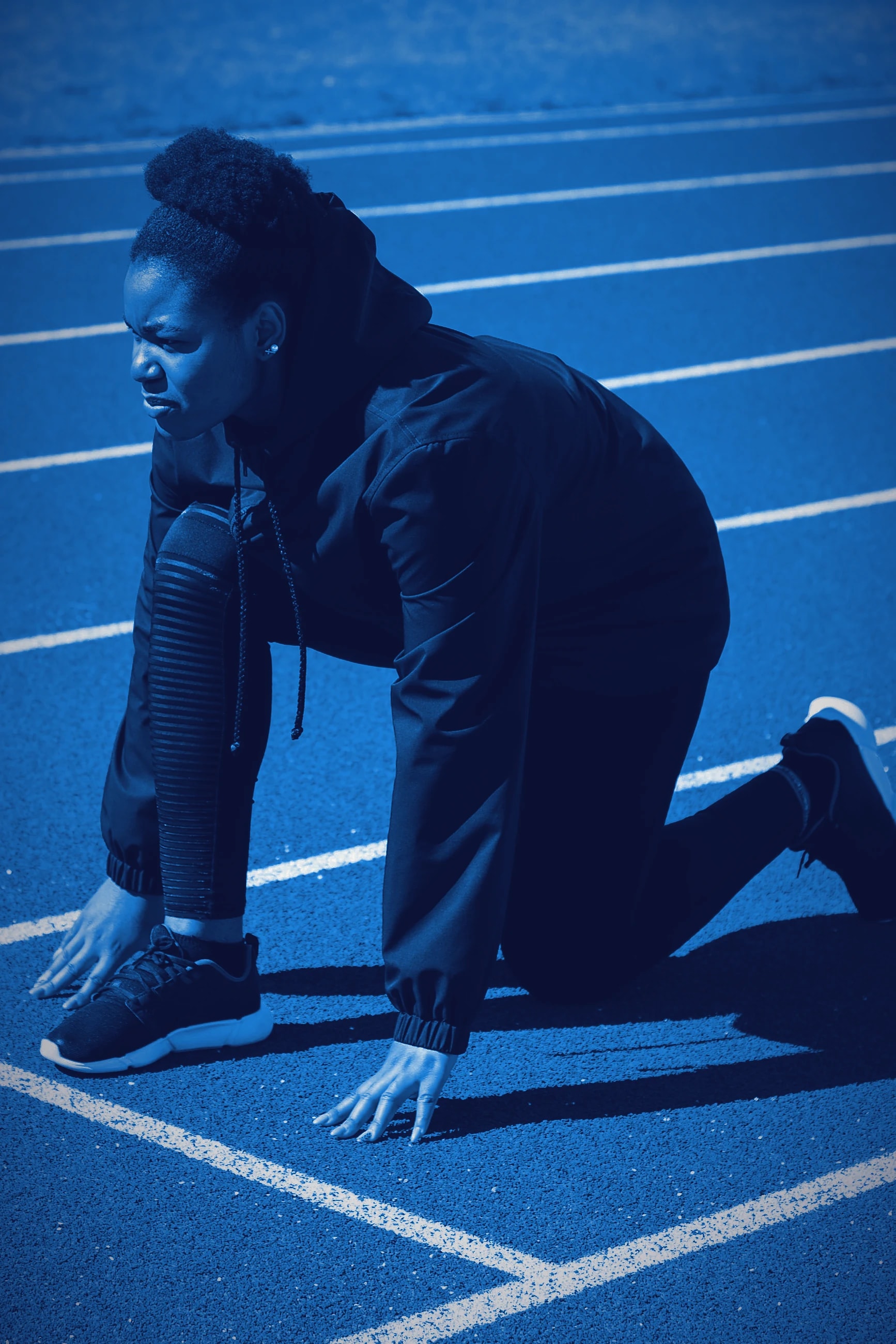 A sprinter crouched in the starting blocks on a dark indoor track, head down.