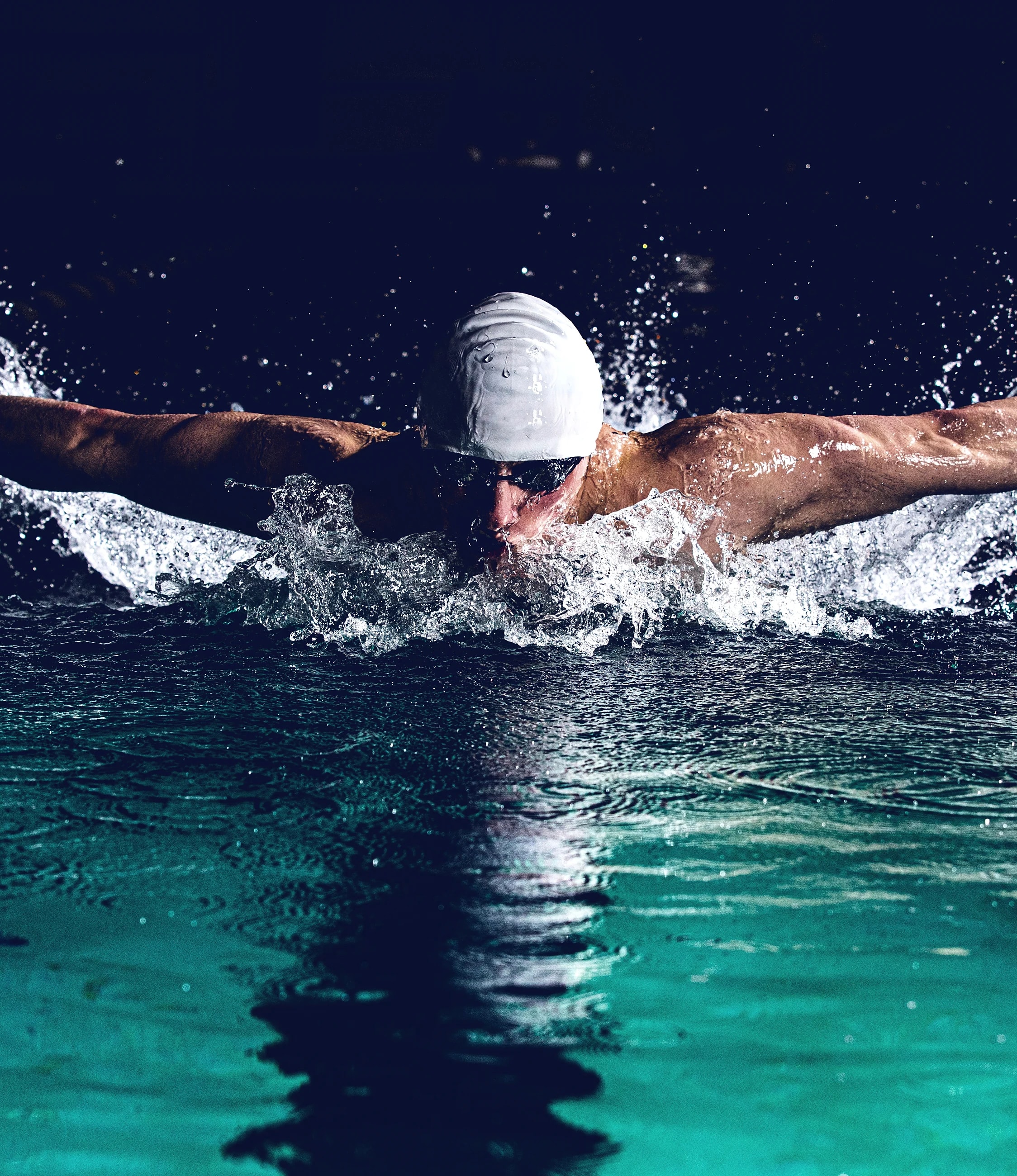 A swimmer mid-butterfly, arms spread wide, throwing a ring of white spray across dark water.