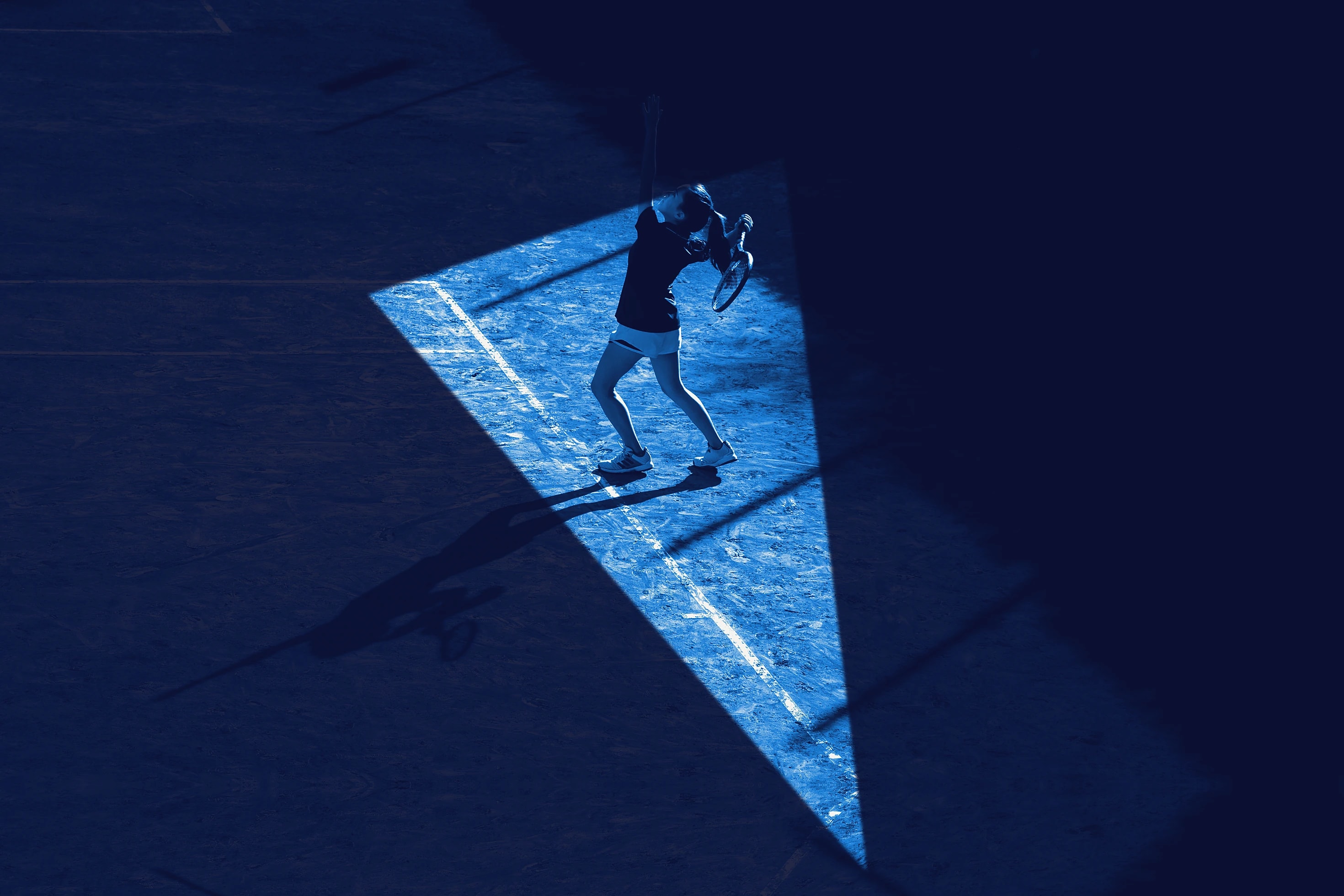 A tennis player at the top of a serve inside a cone of light on a dark court, seen from above.