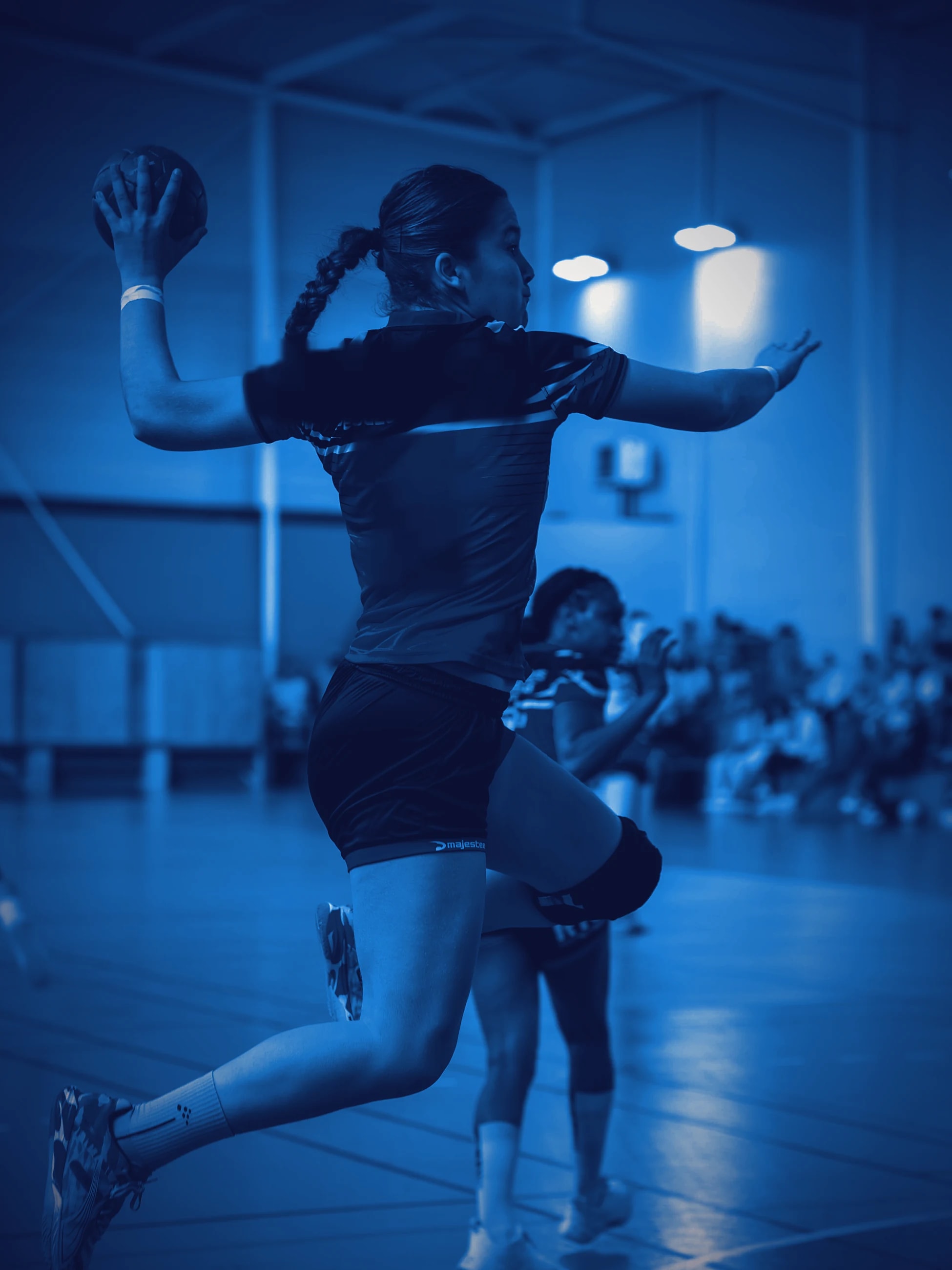 A handball player in mid-air, arm drawn back to shoot, against a dark arena.