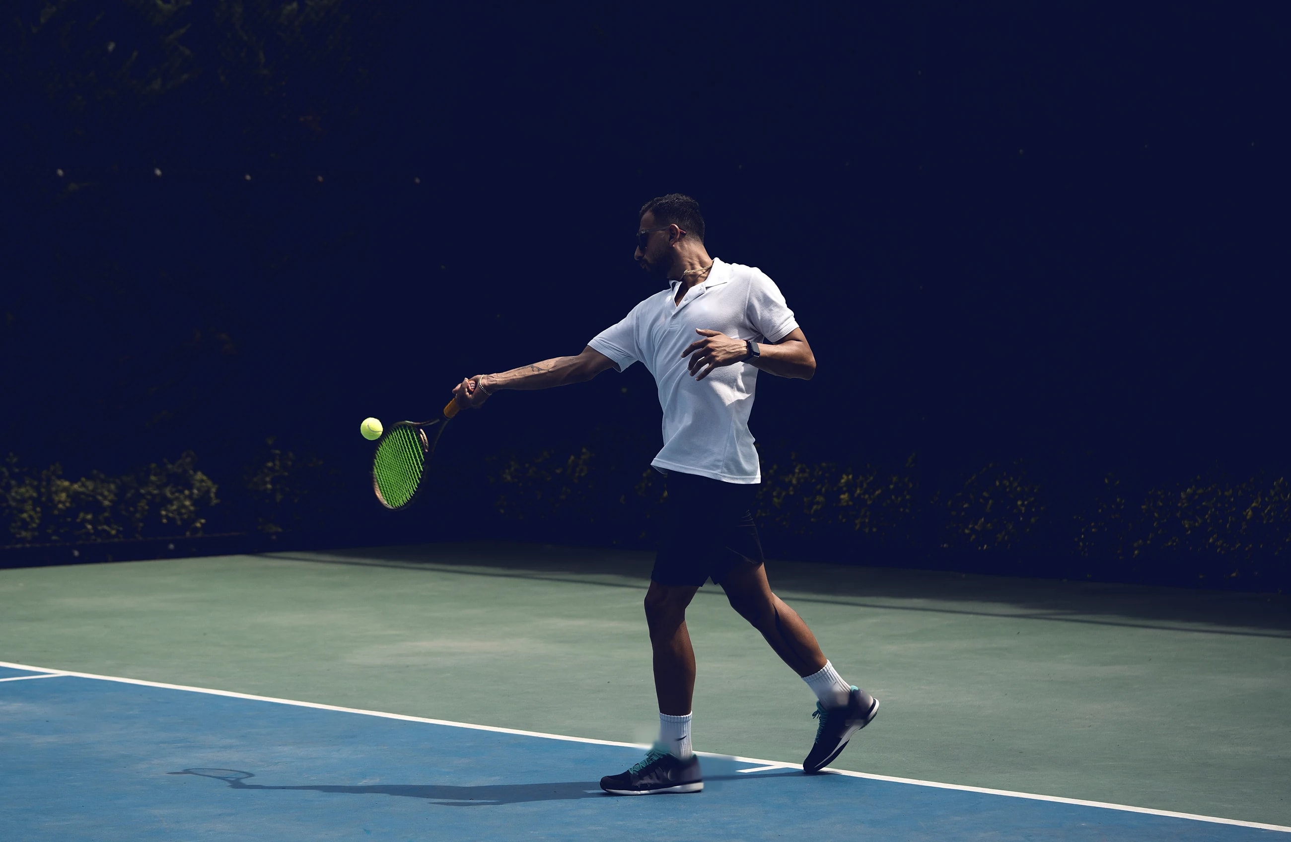 Seen from above, a tennis player in a dark shirt hits a forehand across the white lines of a blue hard court.